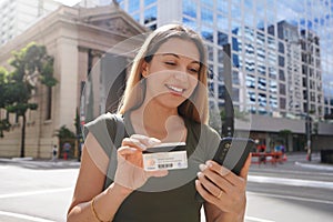 Portrait of young business woman watching her phone holding credit card in finance district avenue