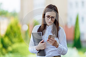 Portrait young business woman wearing white shirt using smartphone out doors. Female reading sms message in working