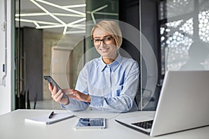 Portrait of a young business woman working in the office at a laptop, using a mobile phone and looking at the camera