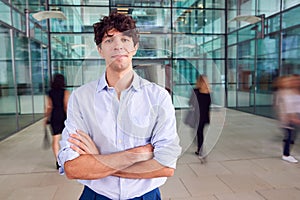 Portrait Of Young Businessman With Crossed Arms Standing In Lobby Of Busy Modern Office