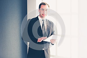 Portrait of young businessman in office with big window. Businessman using tablet computer and looking at camera