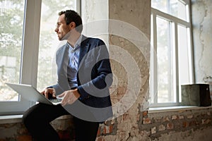 Portrait of young businessman sitting at the window with laptop and working with thoughtful expression. End of business