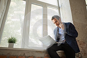 Portrait of young businessman sitting at the window with laptop and working with thoughtful expression. Looking through