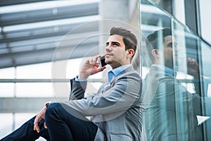 A portrait of young businessman with smartphone sitting in corridor outside office.