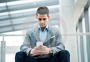 A portrait of young businessman with smartphone sitting in corridor outside office.