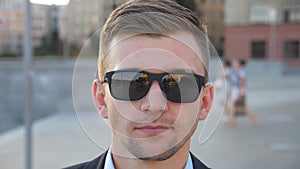 Portrait of young businessman in sunglasses walking along city street. Handsome business man looks at camera strolling