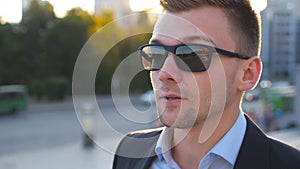 Portrait of young businessman in sunglasses walking along city street with sun flare at background. Handsome business