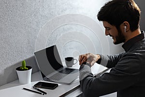 Portrait of young businessman verifies time on smartwatch, using wireless earphones. Laptop, coffee mug, cactus and smartphones.