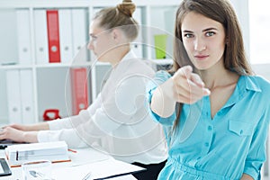 Portrait of young businesswoman pointing straight to the cameraand smiling, with female colleague on the background.