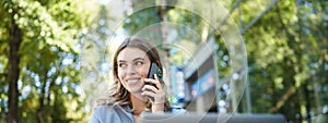 Portrait of young businesswoman sitting outside in park, talking on mobile phone, using laptop. Young woman, student