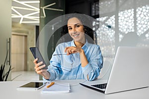 Portrait of young businesswoman at workplace, satisfied smiling woman smiling and looking at camera, holding phone