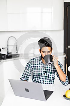 Portrait of a young busy indian man with a beard drinking some coffee and using a laptop computer to work from home
