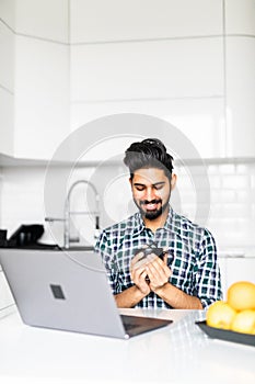 Portrait of a young busy indian man with a beard drinking some coffee and using a laptop computer to work from home