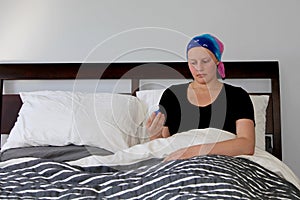 Portrait of a young cancer patient in a headscarf in bed looking at a bottle of pills