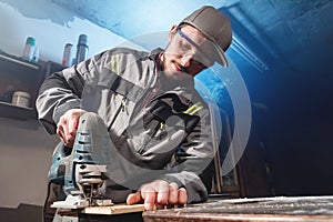 Portrait of a young carpenter joiner with electric jigsaw in the hands of a worker in a home workshop. Starting a