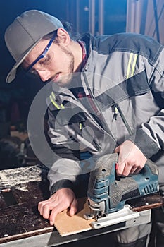 Portrait of a young carpenter joiner with electric jigsaw in the hands of a worker in a home workshop. Starting a