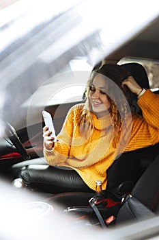 Portrait of a young casual woman in orange sweater texting on her smartphone or making video call while driving or
