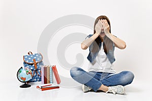 Portrait of young casual woman student with opened mouth covering face with hands, sitting near globe, backpack, school