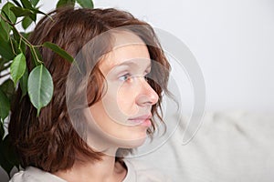 Portrait of a young caucasian brunette girl with curls in a beige dress on a background of a white wall and green leaves