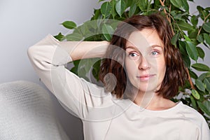 Portrait of a young caucasian brunette girl with curls in a beige dress on a background of a white wall and green leaves