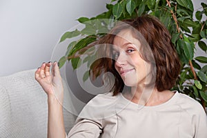 Portrait of a young caucasian brunette girl with curls in a beige dress on a background of a white wall and green leaves