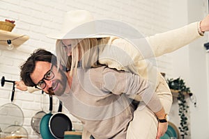 Portrait of young Caucasian couple woman sitting on husband& x27;s back man smiling to the camera kitchen background