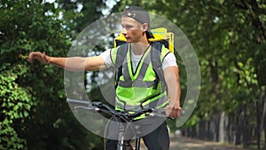 Portrait of young Caucasian delivery man in green vest with yellow backpack riding bicycle gesturing thumb up looking