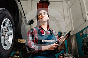 Portrait of a young caucasian female mechanic, in uniform, with a tablet in her hands. The wheel of the car on the right. Bottom