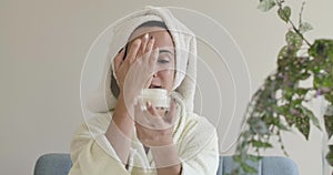 Portrait of a young Caucasian girl in white bathrobe and hair towel applying face cream. Beautiful woman trying