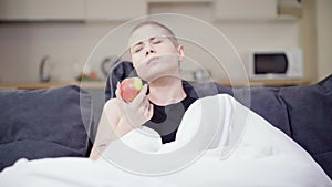 Portrait of young Caucasian hairless woman eating healthful apple sitting on couch at home. Ill female patient