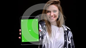 Portrait of young caucasian long-haired girl in shirt smilingly showing green screen of tablet into camera on black photo