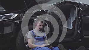 Portrait of young caucasian male auto mechanic in headphones leaning against car wheel and using smartphone