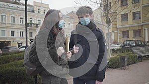 Portrait of young Caucasian male college student in face mask talking to female groupmate outdoors. Boy and girl wearing