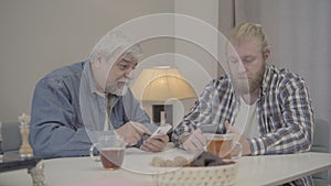 Portrait of young Caucasian man and senior retiree rubbing eyes simultaneously as watching video on smartphones