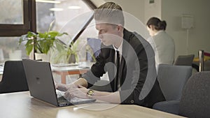 Portrait of young Caucasian man sitting in open space office and typing on laptop keyboard. Stressed male office worker