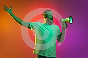 Portrait of young caucasian man speaking into megaphone isolated over gradient orange pink background in neon lights.