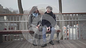 Portrait of young Caucasian man and woman sitting on bench in city park and talking. Happy teenage students dating