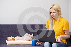 Portrait of Young Caucasian Mother WIth Baby Boy Remote Working Using Laptop and Drinking Tea or Coffee Indoors