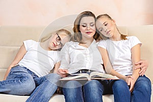 Portrait of Young Caucasian Mother Reading a Book Together with Her Twin Daughters While Sitting on Sofa Indoors