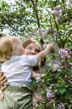 Portrait of young Caucasian mother who is holding her son and touching flowers of lilac tree. Happy family time together