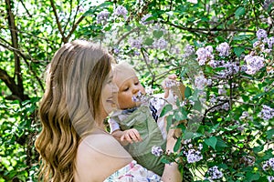 Portrait of young Caucasian mother who is holding her son and touching flowers of lilac tree. Happy family time together