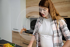 Portrait of a young pregnant woman cooking at home, preparing a healthy vegetable meal, while standing at the kitchen island