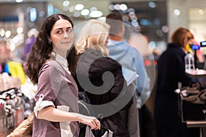 Portrait of young Caucasian smiling woman looking back and standing in store queue to pay for purchase. Concept of