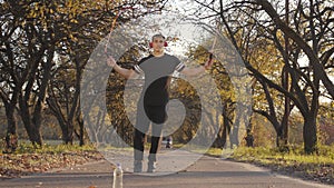 Portrait of a young Caucasian sportsman in headphones and black sportswear jumping rope in the autumn park on sunset