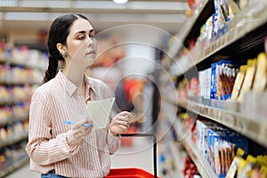 Portrait of young Caucasian woman choosing groceries in supermarket. Millennial with list of products is shopping. In
