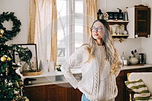 Portrait of young caucasian woman in glasses standing on the kitchen at home with Christmas decoration and looking in window
