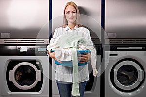 portrait of young caucasian woman holding a basket of clothes to be washed in a automatic laundry
