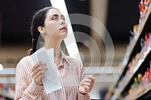 Portrait of young caucasian woman holds products list and choosing food. Showcase and shelves at background. Concept of