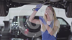 Portrait of young Caucasian woman standing under car and tightening screws. Blond female auto mechanic in blue workrobe