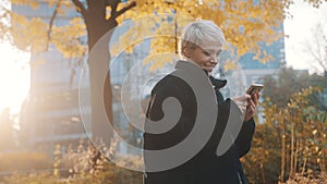 Portrait of young caucasian woman using phone in the city park in front of office building in autumn